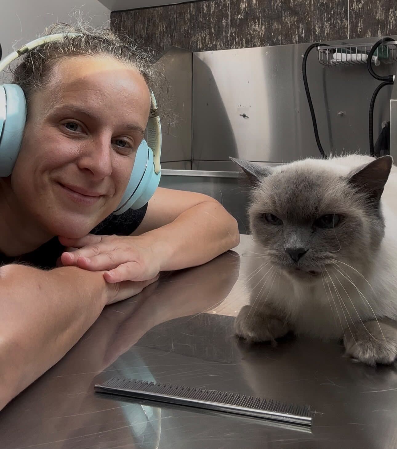 Sandi at the grooming table, chin resting on her hands, with a freshly groomed grey and cream cat sitting beside her.