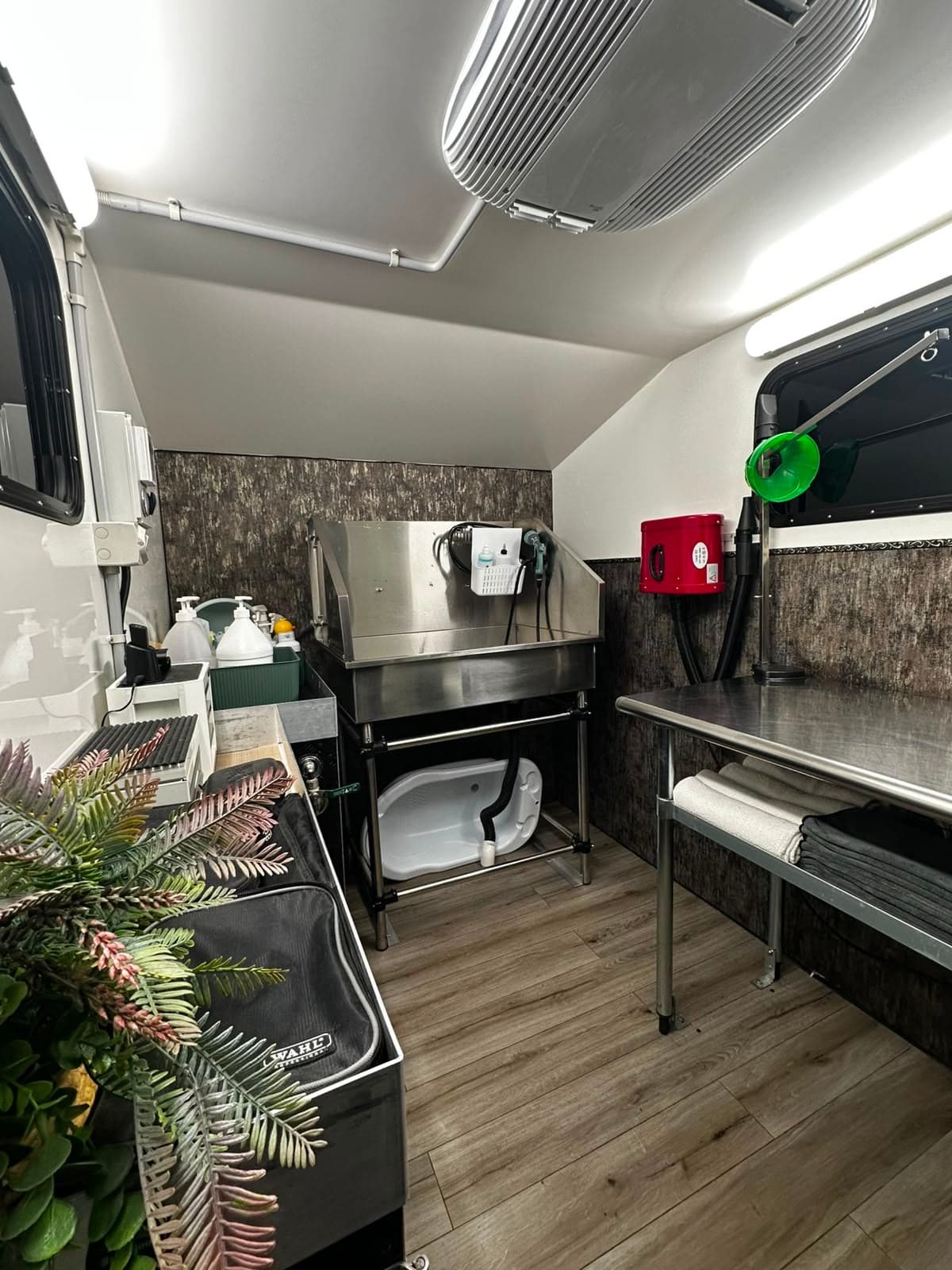 The stainless bath inside the trailer, raised on a frame with a step underneath, a hose and shampoo within reach, and the grooming table alongside it stacked with folded towels.