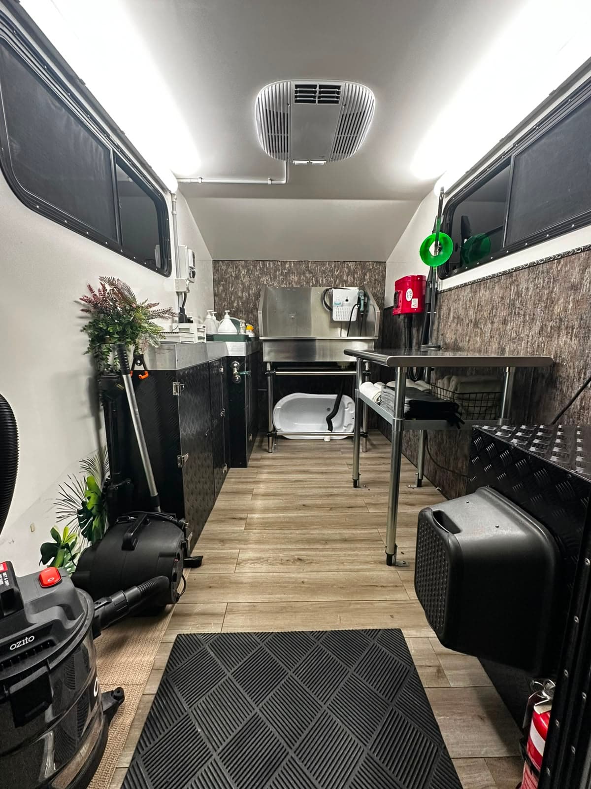 Inside the grooming trailer: a stainless steel bath at the far end with a hose and dryer beside it, a stainless grooming table down one wall with towels folded on the shelf underneath, timber-look flooring and a rubber mat at the door.